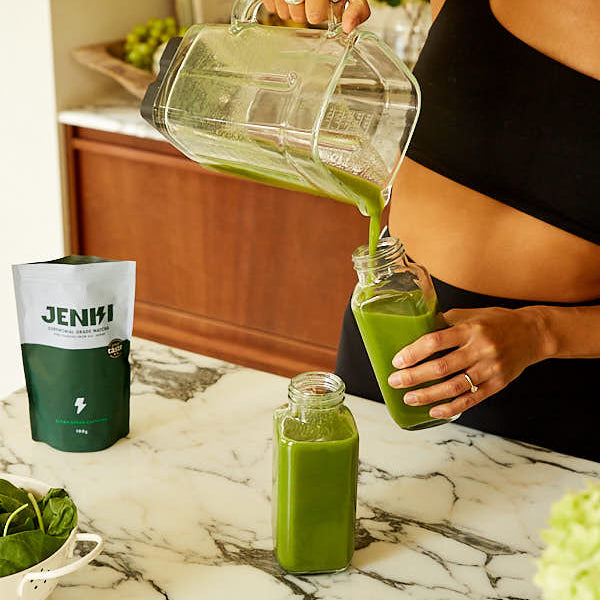 Person pouring green juice from a pitcher into a glass jar on a marble countertop.