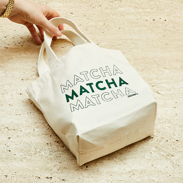 White tote bag with 'MATCHA MATCHA MATCHA' text held by a hand on a wooden surface