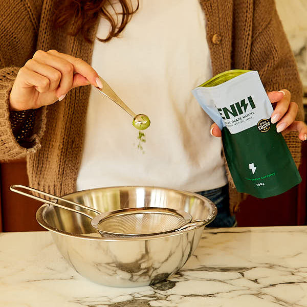 Person holding a package of JENKI matcha and using a spoon with a green substance over a metal strainer on a marble surface.